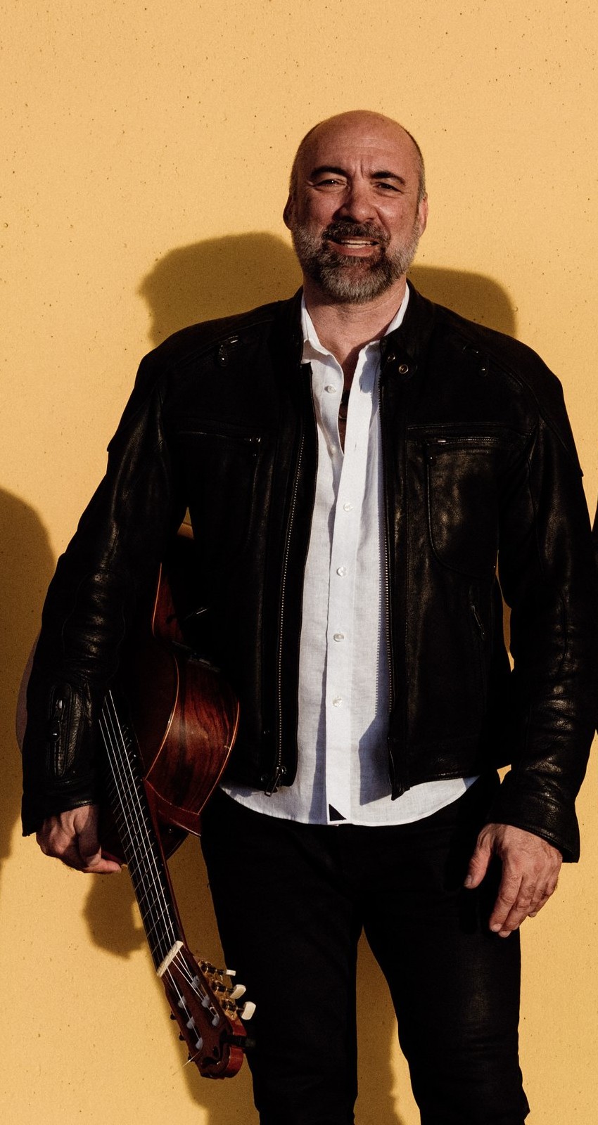 Petar Jankovic against a warm yellow wall, leather jacket, guitar at his side